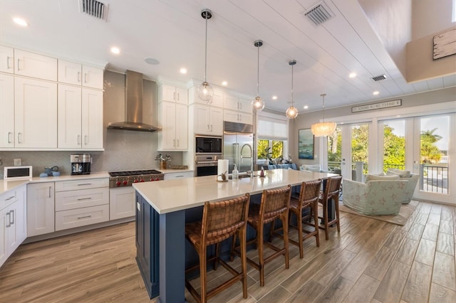 kitchen with decorative backsplash, built in appliances, wall chimney exhaust hood, an island with sink, and light hardwood / wood-style floors