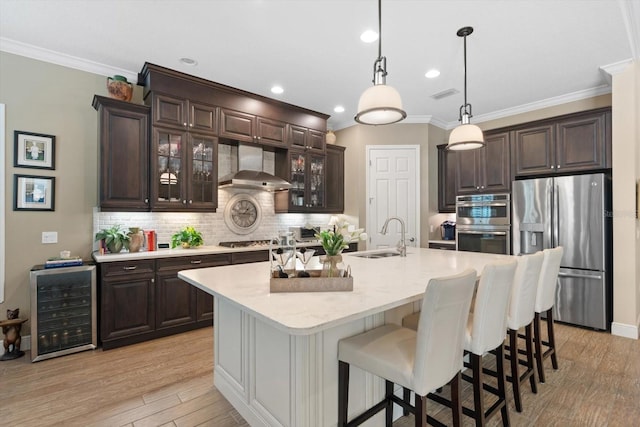 kitchen with appliances with stainless steel finishes, sink, wall chimney range hood, wine cooler, and a kitchen island with sink