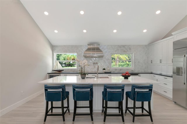 kitchen featuring stainless steel built in fridge, a center island with sink, tasteful backsplash, wall chimney exhaust hood, and white cabinets