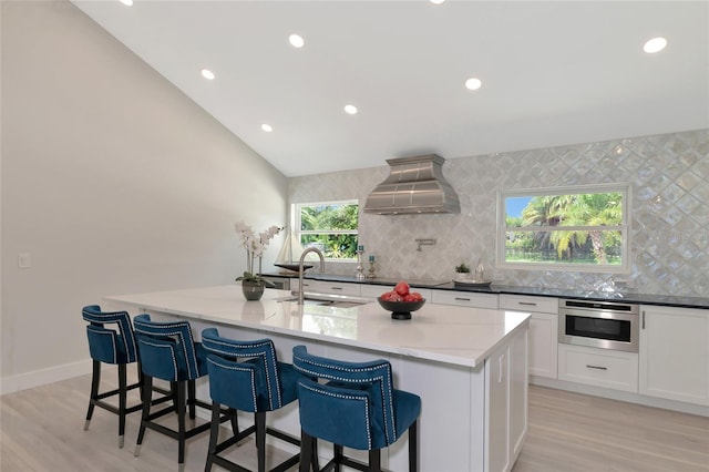 kitchen with custom exhaust hood, dark stone counters, an island with sink, oven, and a sink