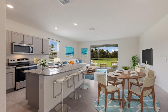 kitchen with sink, an island with sink, gray cabinetry, light tile patterned floors, and stainless steel appliances