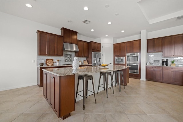 kitchen featuring stainless steel appliances, tasteful backsplash, a kitchen island with sink, a kitchen breakfast bar, and light stone countertops