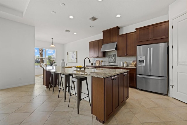 kitchen with light stone counters, stainless steel fridge, pendant lighting, a breakfast bar area, and a kitchen island with sink