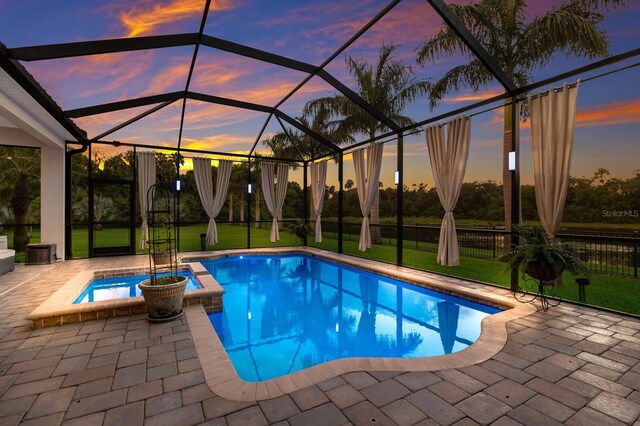 pool at dusk featuring glass enclosure, an in ground hot tub, and a patio area