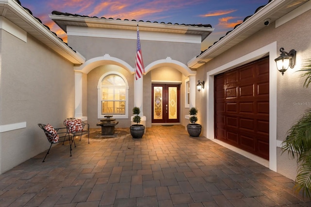 exterior entry at dusk with a garage, french doors, and stucco siding