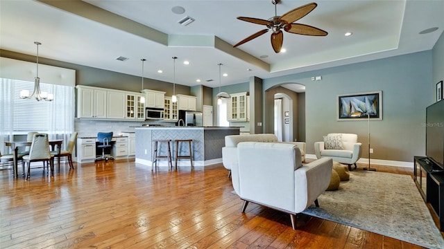 living room with a tray ceiling, light wood-style flooring, ceiling fan with notable chandelier, and arched walkways