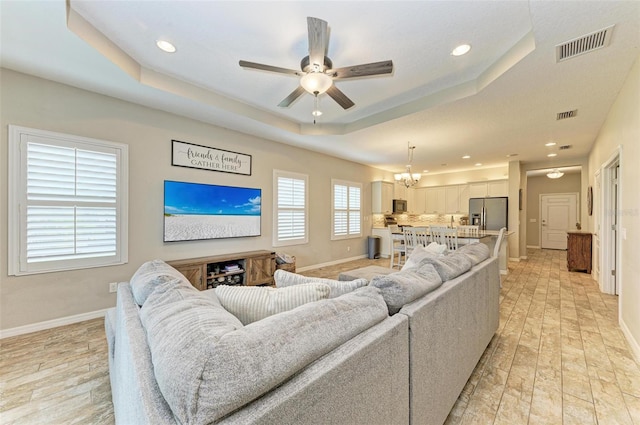 living room with ceiling fan with notable chandelier, a tray ceiling, and light hardwood / wood-style floors