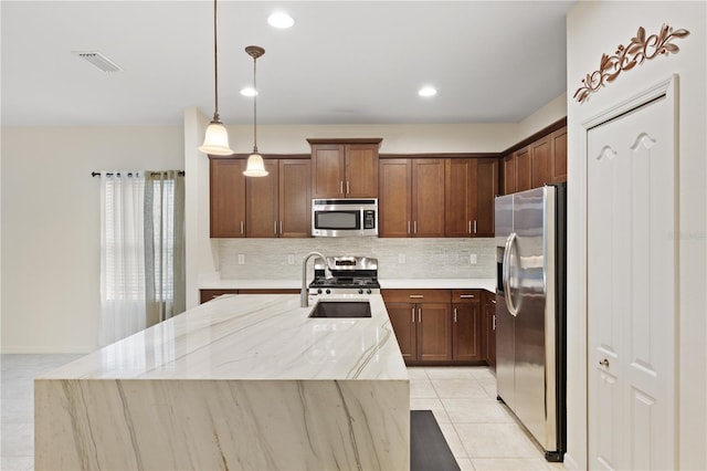 kitchen featuring light stone counters, backsplash, an island with sink, stainless steel appliances, and decorative light fixtures