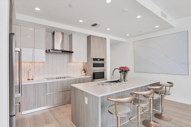 kitchen featuring sink, wall chimney exhaust hood, appliances with stainless steel finishes, light hardwood / wood-style floors, and a kitchen bar