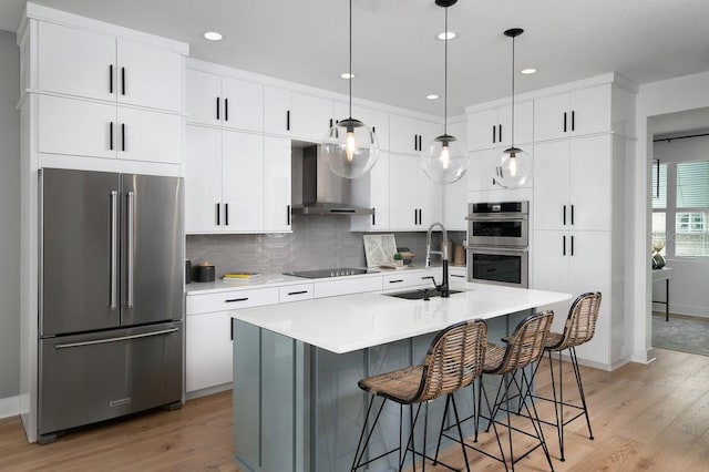 kitchen with appliances with stainless steel finishes, wall chimney exhaust hood, sink, an island with sink, and light wood-type flooring