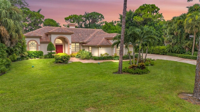 mediterranean / spanish-style home with driveway, a tiled roof, a lawn, stucco siding, and a chimney