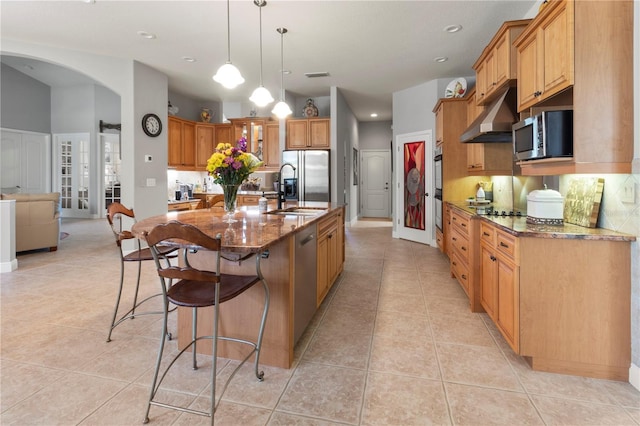 kitchen with light tile patterned flooring, under cabinet range hood, a sink, appliances with stainless steel finishes, and dark stone counters