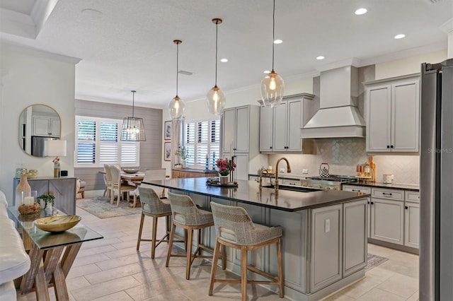 kitchen featuring custom range hood, gray cabinets, a kitchen breakfast bar, a center island with sink, and ornamental molding