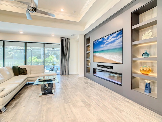 living room with light wood-type flooring, a raised ceiling, built in features, ceiling fan, and ornamental molding