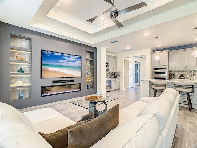 living room with light hardwood / wood-style flooring, built in shelves, a tray ceiling, ceiling fan, and ornamental molding