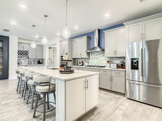 kitchen with appliances with stainless steel finishes, wall chimney exhaust hood, decorative backsplash, a kitchen island with sink, and a breakfast bar