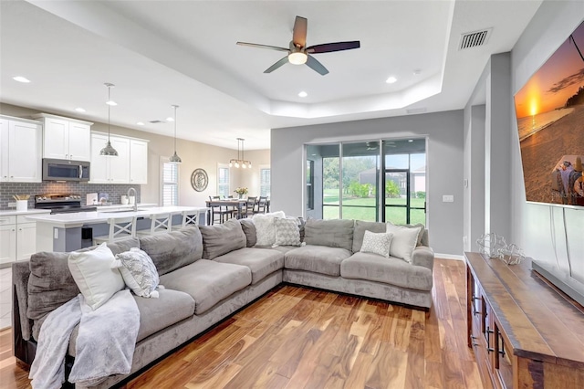 living room with a raised ceiling, ceiling fan, sink, and light wood-type flooring