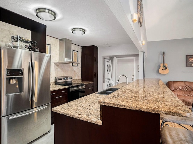 kitchen with wall chimney exhaust hood, stacked washing maching and dryer, stainless steel appliances, and dark brown cabinets