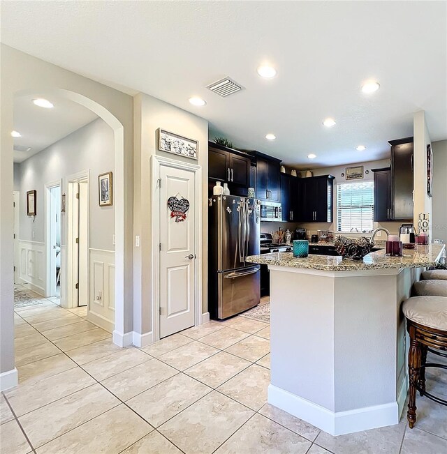 kitchen with light tile patterned floors, light stone countertops, stainless steel appliances, kitchen peninsula, and a kitchen bar