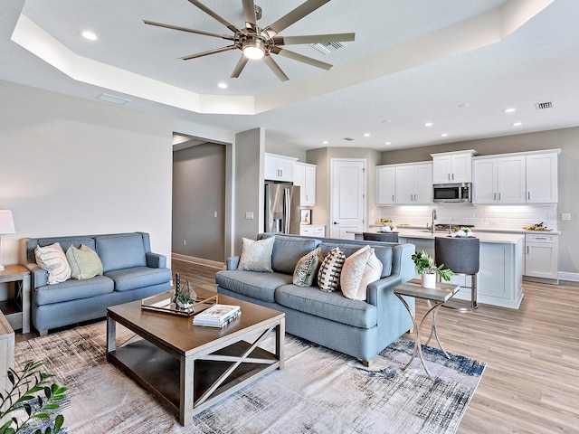 living room with a textured ceiling, a raised ceiling, light hardwood / wood-style flooring, sink, and ceiling fan