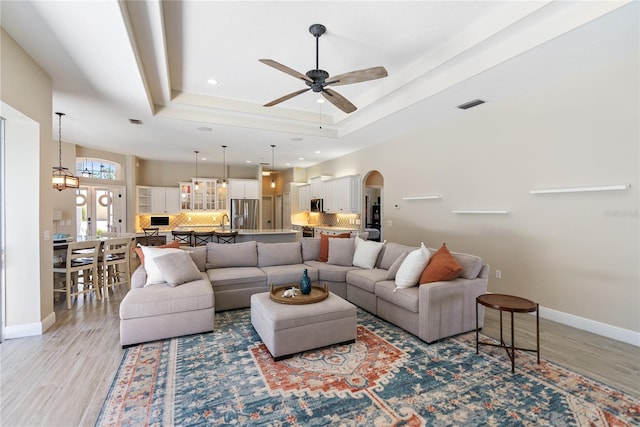 living room featuring french doors, ceiling fan with notable chandelier, light hardwood / wood-style floors, and a raised ceiling