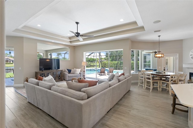 living room featuring a fireplace, ceiling fan with notable chandelier, a tray ceiling, and light hardwood / wood-style flooring