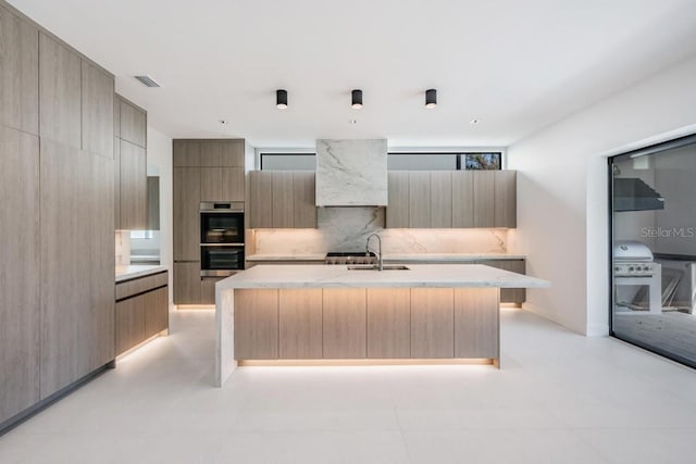 kitchen with sink, light brown cabinets, a center island with sink, and stainless steel double oven