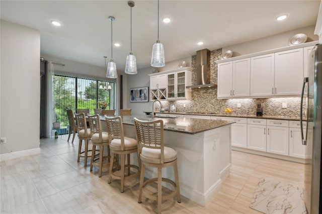 kitchen featuring wall chimney range hood, dark stone countertops, an island with sink, and white cabinetry