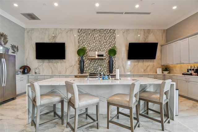 kitchen featuring light stone countertops, stainless steel fridge, a kitchen breakfast bar, decorative backsplash, and a center island with sink