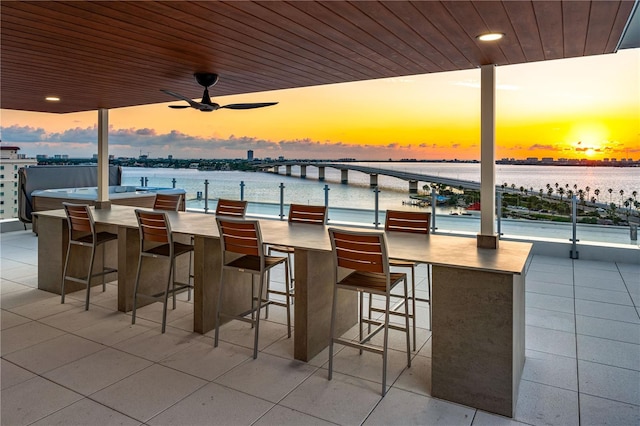 patio terrace at dusk featuring ceiling fan, a water view, and an outdoor bar