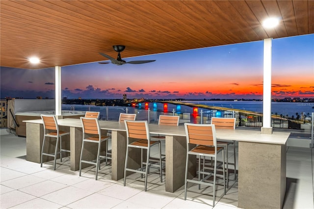 patio terrace at dusk with an outdoor bar, ceiling fan, and a water view