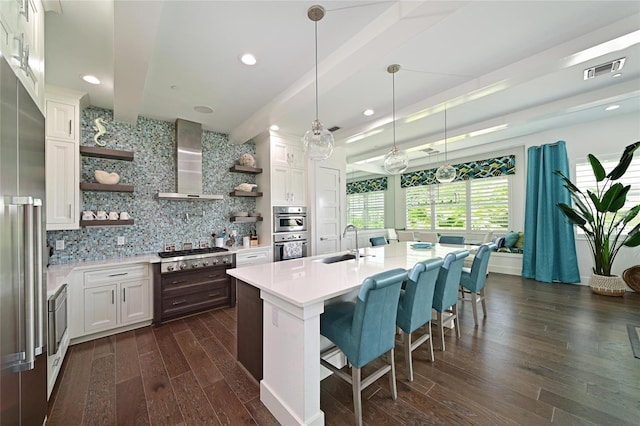 kitchen with wall chimney range hood, appliances with stainless steel finishes, a kitchen island with sink, white cabinets, and decorative light fixtures