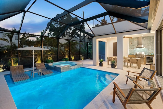 pool at dusk with an in ground hot tub, area for grilling, exterior kitchen, glass enclosure, and a patio area