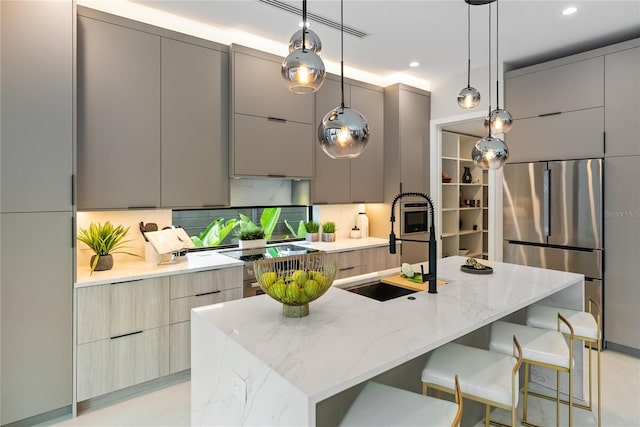 kitchen featuring sink, gray cabinetry, a center island with sink, hanging light fixtures, and stainless steel refrigerator