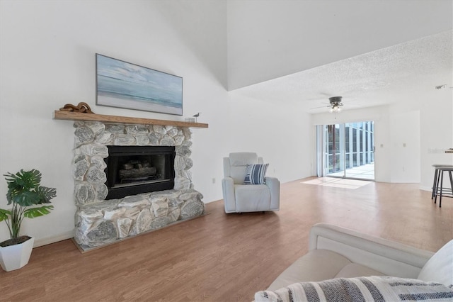 living area featuring a ceiling fan, a stone fireplace, a textured ceiling, and wood finished floors