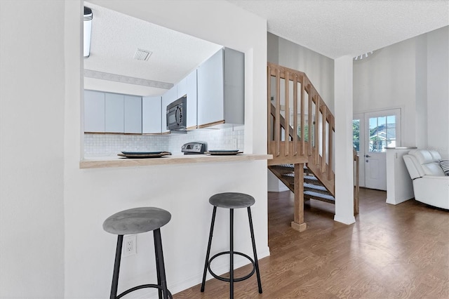 kitchen featuring backsplash, a textured ceiling, wood finished floors, black microwave, and a kitchen breakfast bar