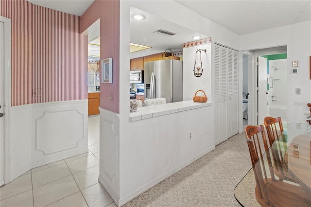 kitchen featuring a wainscoted wall, light tile patterned floors, brown cabinets, a peninsula, and stainless steel fridge