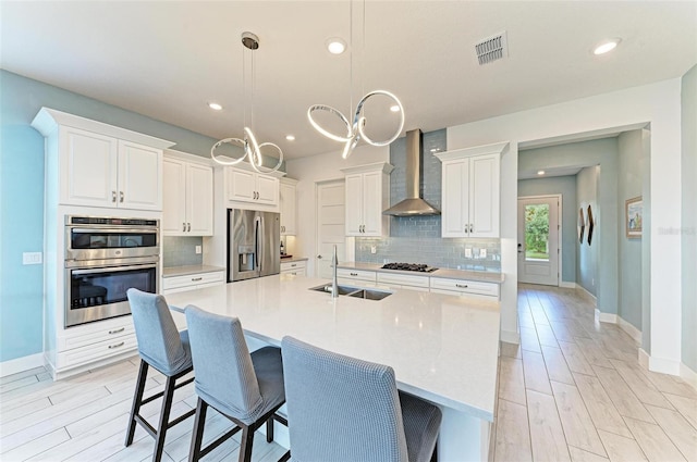 kitchen with pendant lighting, an island with sink, sink, appliances with stainless steel finishes, and wall chimney range hood