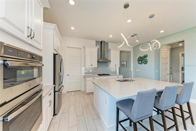 kitchen with white cabinets, stainless steel appliances, sink, wall chimney exhaust hood, and a center island with sink