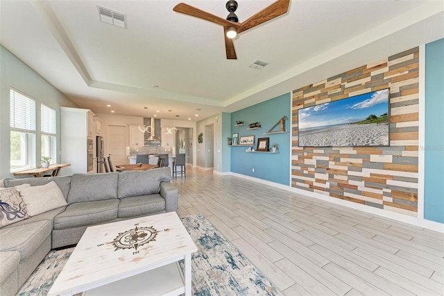 living room with ceiling fan, wood walls, light wood-type flooring, and a tray ceiling