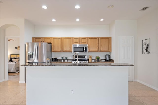 kitchen featuring light tile patterned floors, stainless steel appliances, dark stone countertops, and a kitchen island with sink