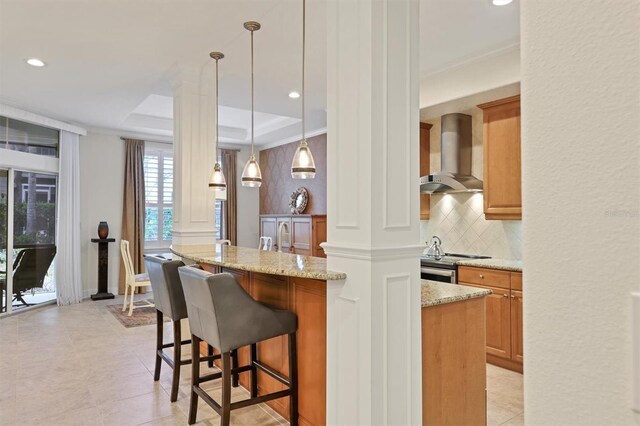 kitchen with light stone counters, a kitchen breakfast bar, ornate columns, and wall chimney range hood