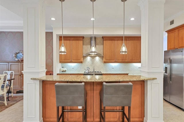 kitchen with wall chimney range hood, stainless steel refrigerator with ice dispenser, a breakfast bar, light stone countertops, and ornate columns