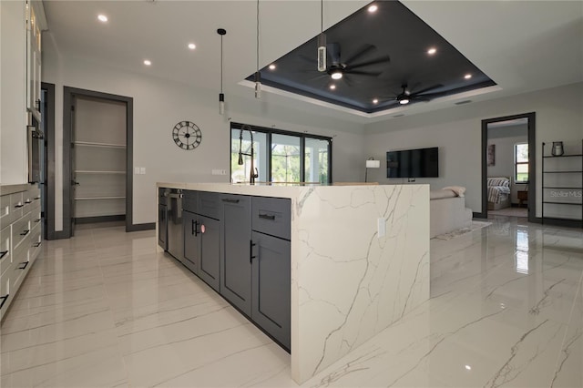 kitchen with hanging light fixtures, light stone countertops, a tray ceiling, an island with sink, and ceiling fan