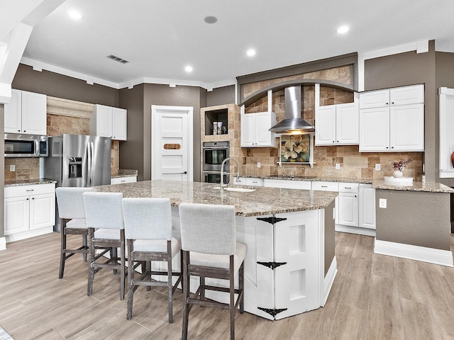 kitchen with wall chimney range hood, stainless steel appliances, a center island with sink, and white cabinetry