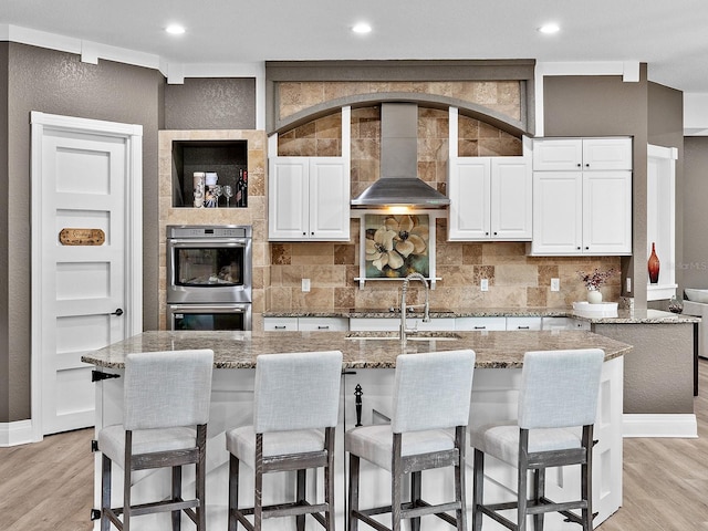 kitchen with a kitchen island with sink, wall chimney exhaust hood, and white cabinets