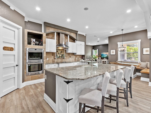 kitchen featuring hanging light fixtures, light wood-type flooring, white cabinetry, wall chimney range hood, and a breakfast bar