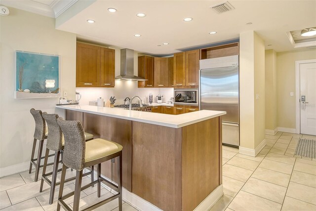 kitchen featuring light tile patterned floors, crown molding, wall chimney exhaust hood, kitchen peninsula, and built in appliances