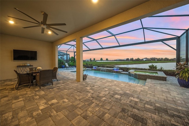 pool at dusk featuring ceiling fan, a lanai, an in ground hot tub, a water view, and a patio