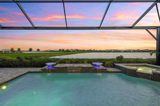 pool at dusk featuring a lanai, an in ground hot tub, a water view, and pool water feature